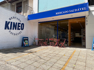 Fachada branca e azul da KINEO, com a entrada da loja e mesas vermelhas na calçada.